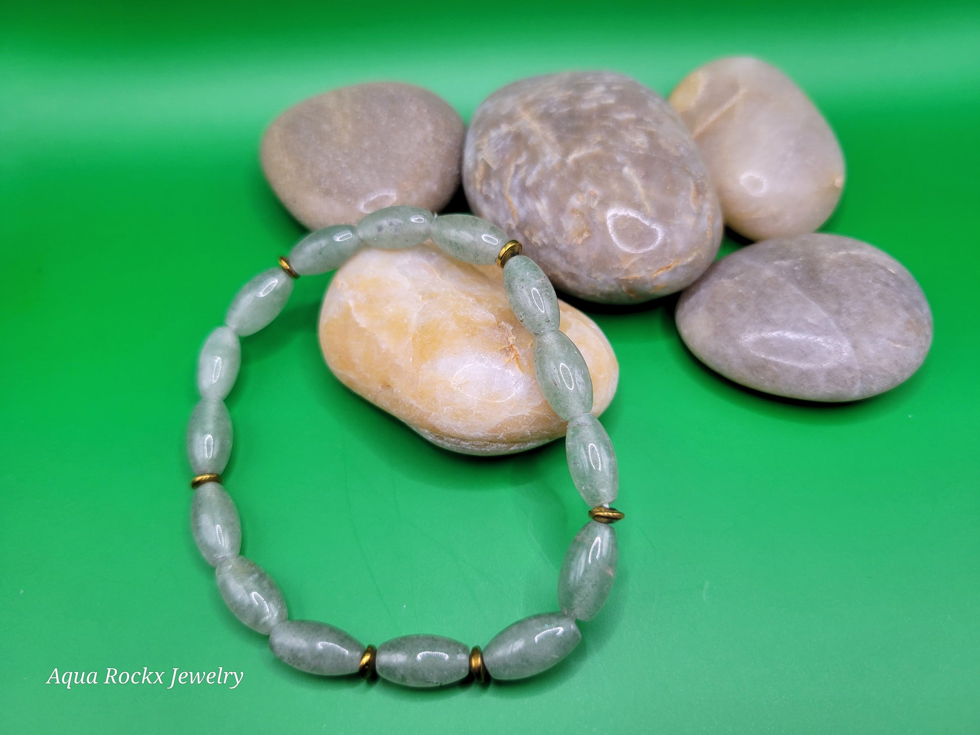 Green beaded bracelet with gold accents on pebbles against a green background, branded 'Aqua Rockx Jewelry'.