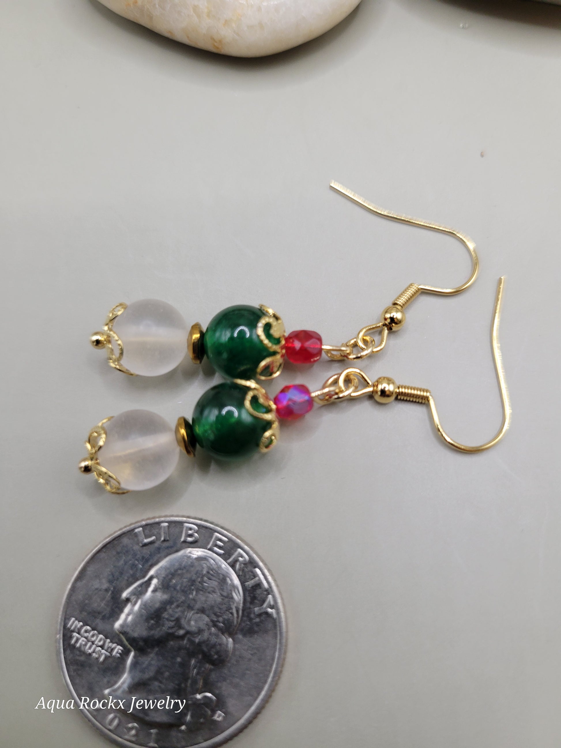 Gold earrings with green and red beads next to a quarter for scale on a light gray background