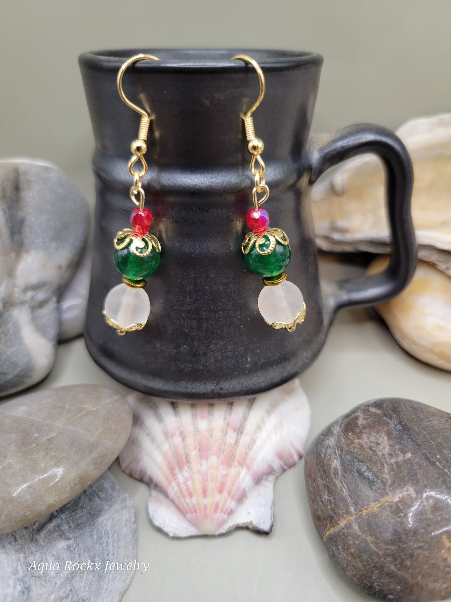 Pair of gold earrings with colorful beads on a black mug surrounded by stones and shells.