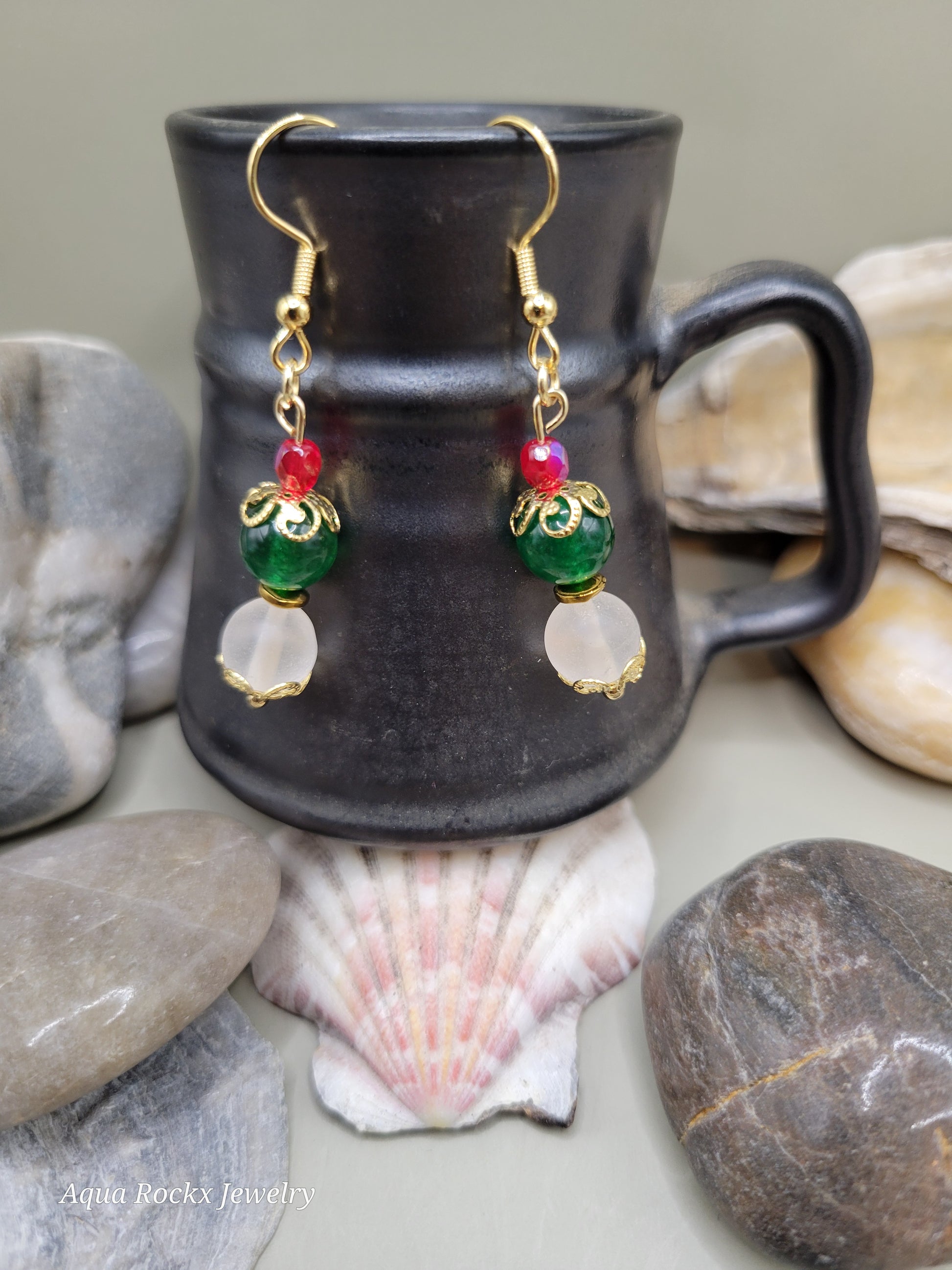 Pair of gold earrings with colorful beads on a black mug surrounded by stones and shells.
